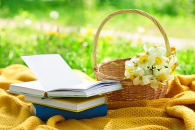 Photo of Stacked books, blanket and wicker basket with daffodil flowers on green grass outdoors