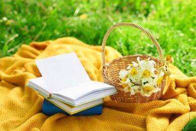 Photo of Stacked books, blanket and wicker basket with daffodil flowers on green grass outdoors