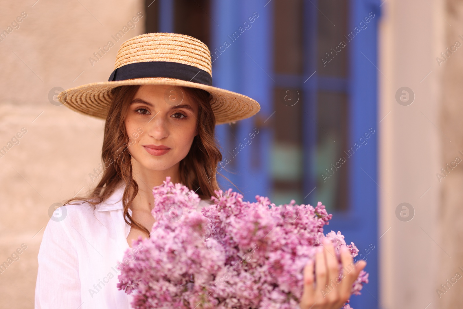 Beautiful woman in hat with bouquet of lilac flowers near building outdoors Photo of Beautiful woman in hat with bouquet of lilac flowers near building outdoors