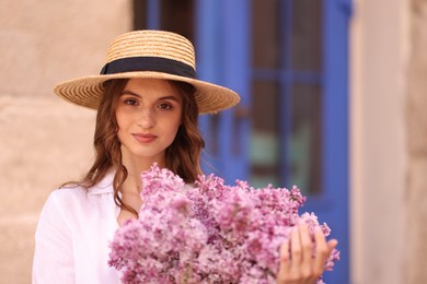 Photo of Beautiful woman in hat with bouquet of lilac flowers near building outdoors