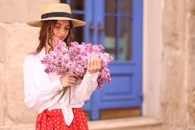 Photo of Beautiful woman in hat with bouquet of lilac flowers near building outdoors. Space for text
