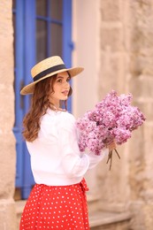 Photo of Beautiful woman in hat with bouquet of lilac flowers near building outdoors