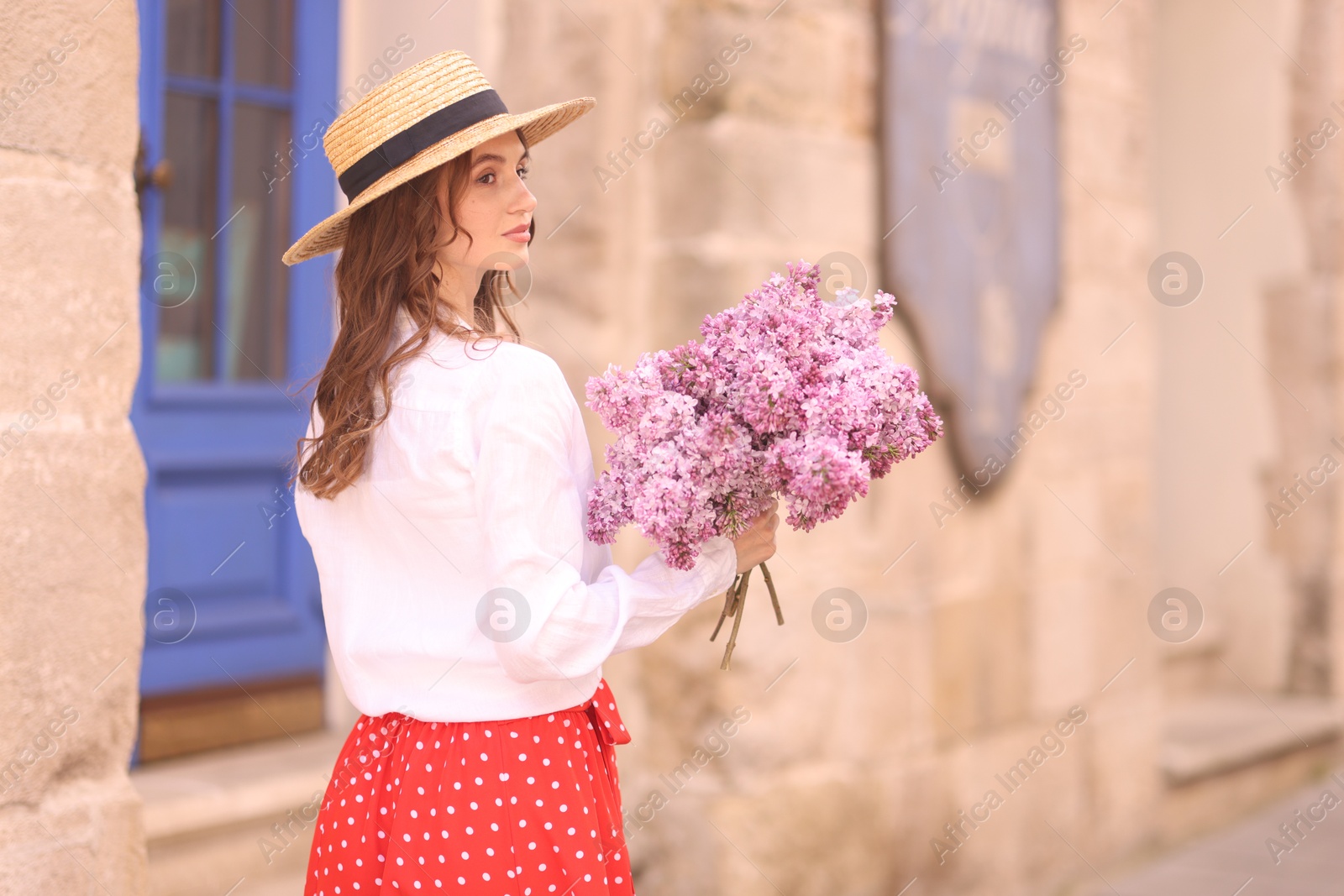 Beautiful woman in hat with bouquet of lilac flowers near building outdoors. Space for text Photo of Beautiful woman in hat with bouquet of lilac flowers near building outdoors. Space for text