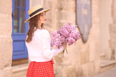 Photo of Beautiful woman in hat with bouquet of lilac flowers near building outdoors. Space for text