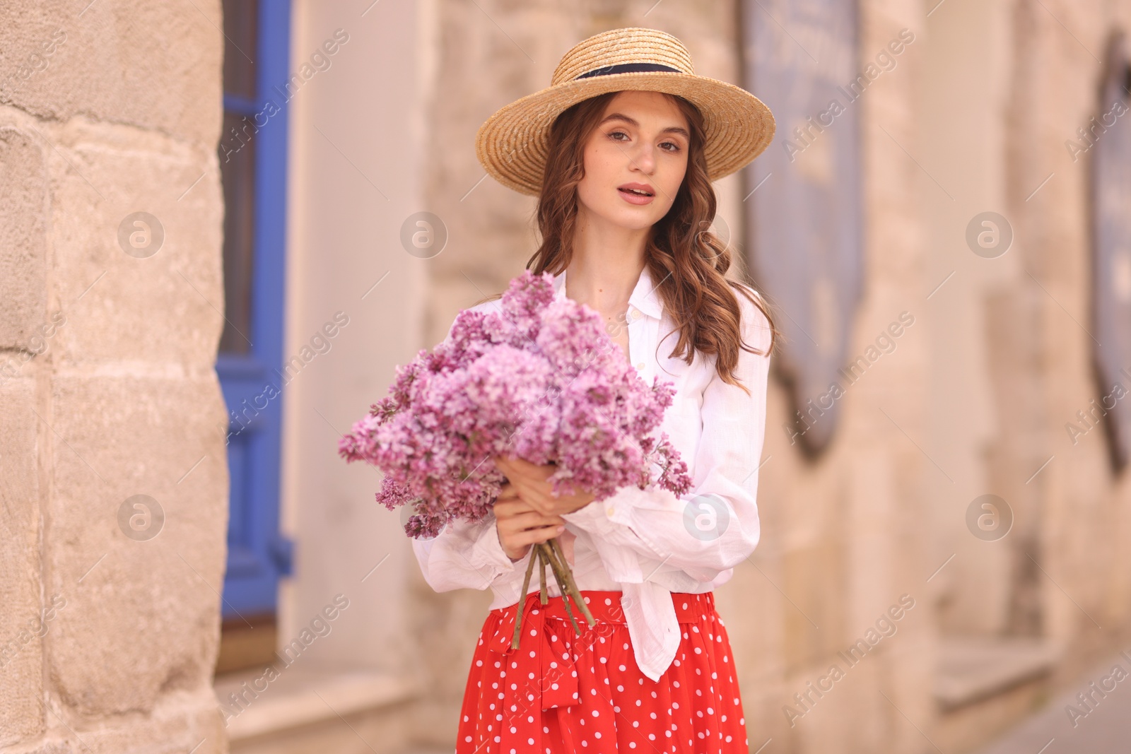 Beautiful woman in hat with bouquet of lilac flowers near building outdoors Photo of Beautiful woman in hat with bouquet of lilac flowers near building outdoors