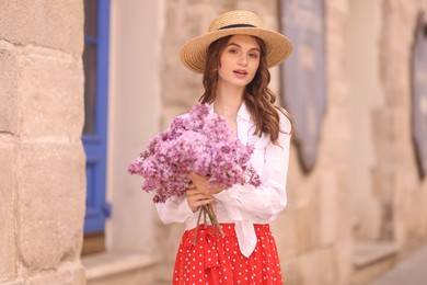 Photo of Beautiful woman in hat with bouquet of lilac flowers near building outdoors