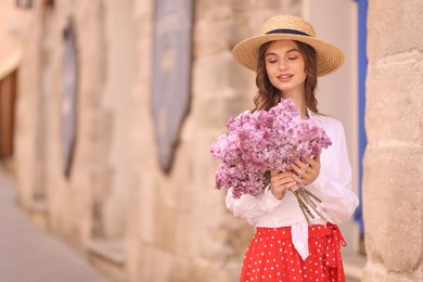 Photo of Smiling woman in hat with bouquet of lilac flowers near building outdoors. Space for text