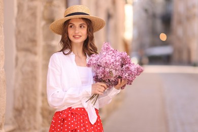 Photo of Smiling woman in hat with bouquet of lilac flowers near building outdoors