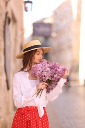 Photo of Beautiful woman in hat with bouquet of lilac flowers near building outdoors