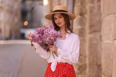 Photo of Beautiful woman in hat with bouquet of lilac flowers near building outdoors