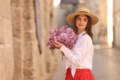 Photo of Beautiful woman in hat with bouquet of lilac flowers near building outdoors