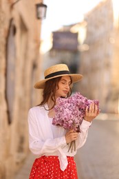 Photo of Beautiful woman in hat with bouquet of lilac flowers near building outdoors
