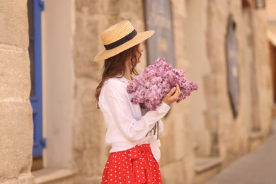 Photo of Woman in hat with bouquet of lilac flowers near building outdoors