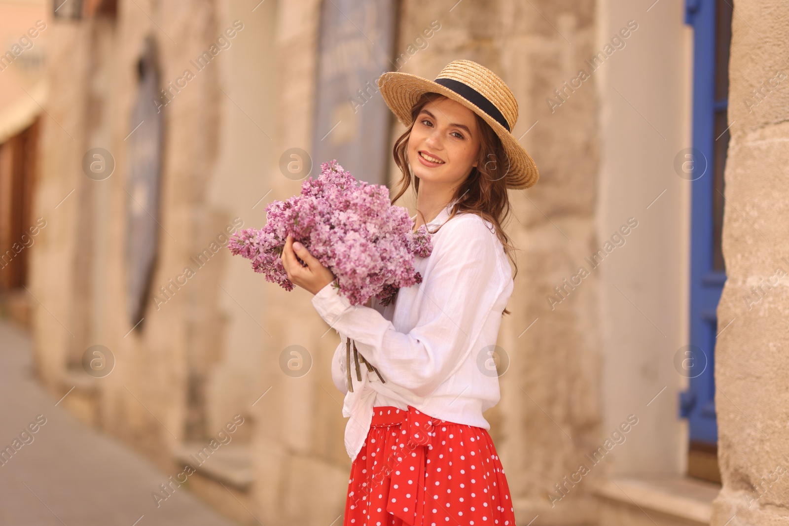 Smiling woman in hat with bouquet of lilac flowers near building outdoors Photo of Smiling woman in hat with bouquet of lilac flowers near building outdoors