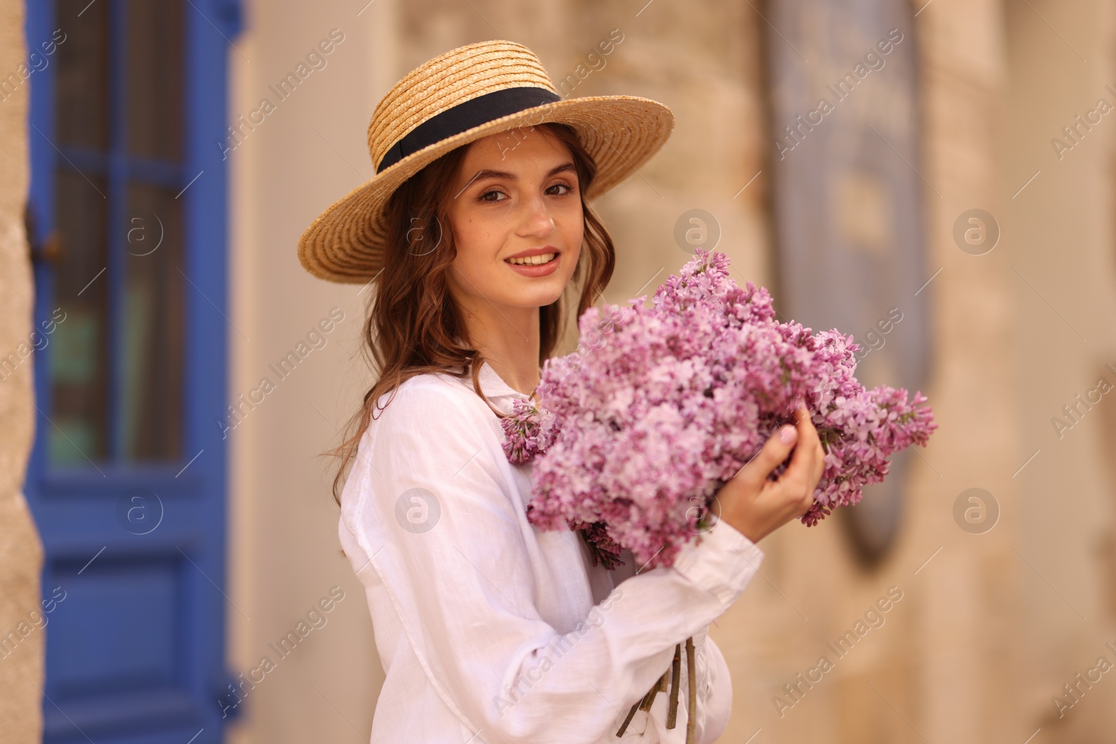 Smiling woman in hat with bouquet of lilac flowers near building outdoors Photo of Smiling woman in hat with bouquet of lilac flowers near building outdoors