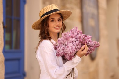Photo of Smiling woman in hat with bouquet of lilac flowers near building outdoors