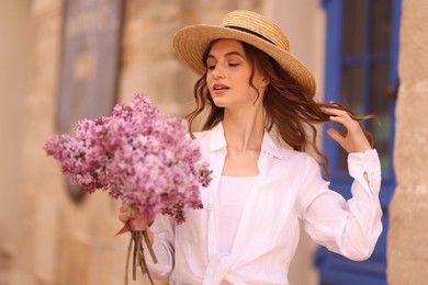 Photo of Beautiful woman in hat with bouquet of lilac flowers near building outdoors