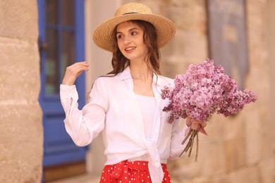 Photo of Smiling woman in hat with bouquet of lilac flowers near building outdoors