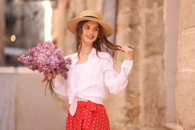 Photo of Smiling woman in hat with bouquet of lilac flowers near building outdoors
