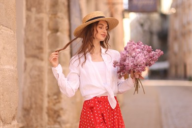 Photo of Beautiful woman in hat with bouquet of lilac flowers near building outdoors