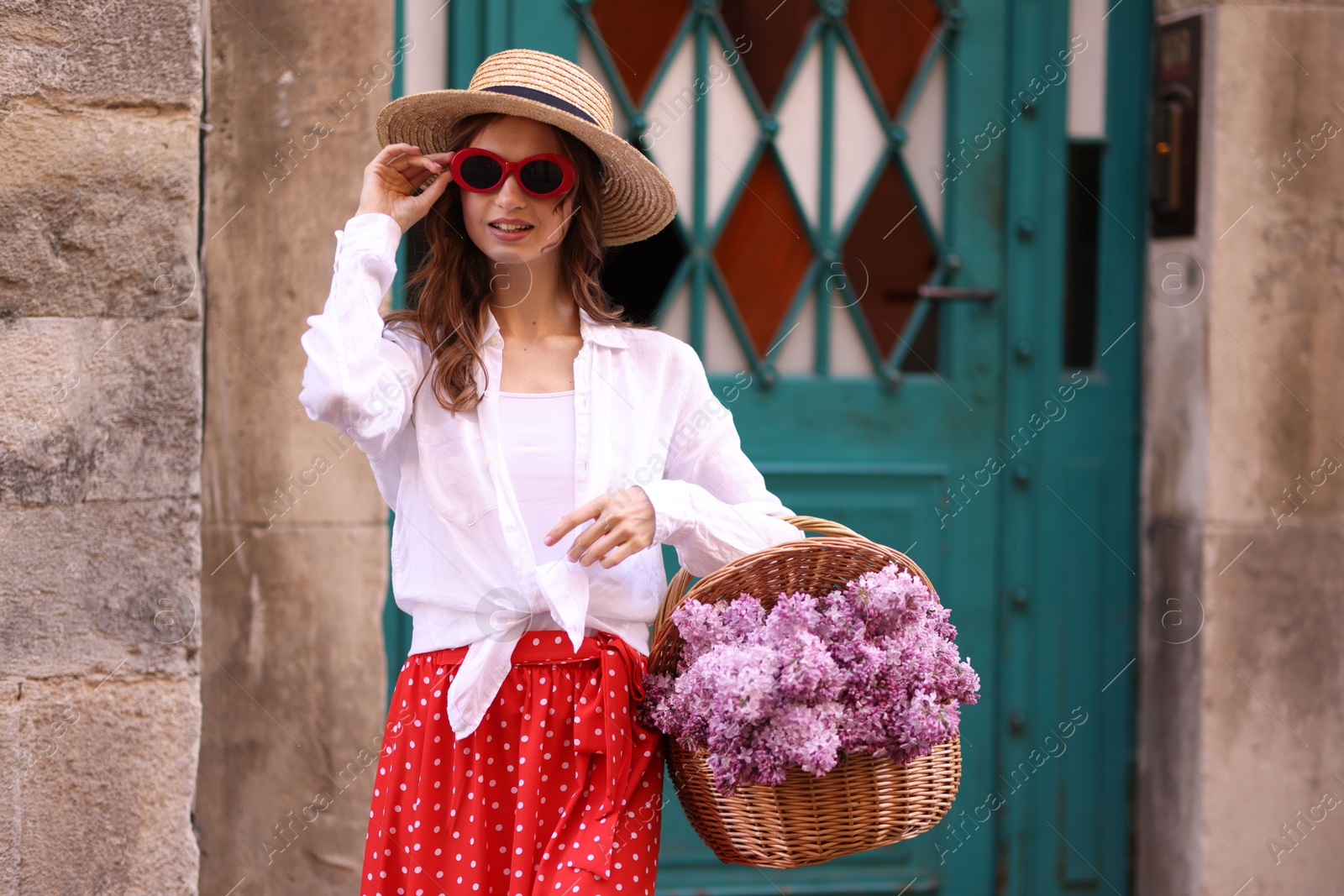 Smiling woman in hat and sunglasses with basket of lilac flowers near building outdoors Photo of Smiling woman in hat and sunglasses with basket of lilac flowers near building outdoors