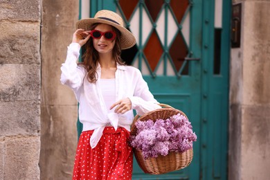 Photo of Smiling woman in hat and sunglasses with basket of lilac flowers near building outdoors