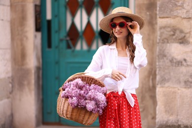 Photo of Smiling woman in hat and sunglasses with basket of lilac flowers near building outdoors
