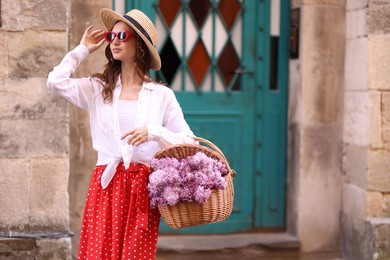 Photo of Beautiful woman in hat and sunglasses with basket of lilac flowers near building outdoors. Space for text