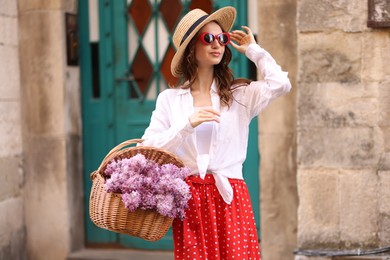 Photo of Beautiful woman in hat and sunglasses with basket of lilac flowers near building outdoors