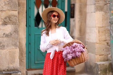 Photo of Smiling woman in hat and sunglasses with basket of lilac flowers near building outdoors