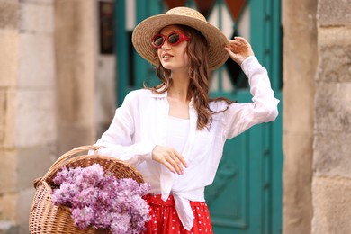 Photo of Smiling woman in hat and sunglasses with basket of lilac flowers near building outdoors