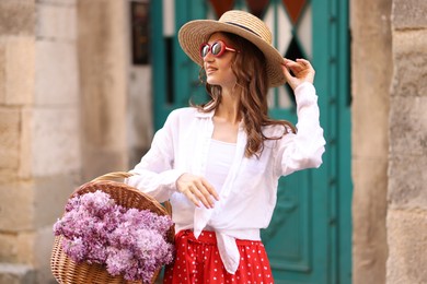 Photo of Smiling woman in hat and sunglasses with basket of lilac flowers near building outdoors