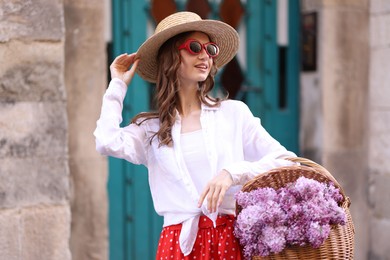 Photo of Smiling woman in hat and sunglasses with basket of lilac flowers near building outdoors