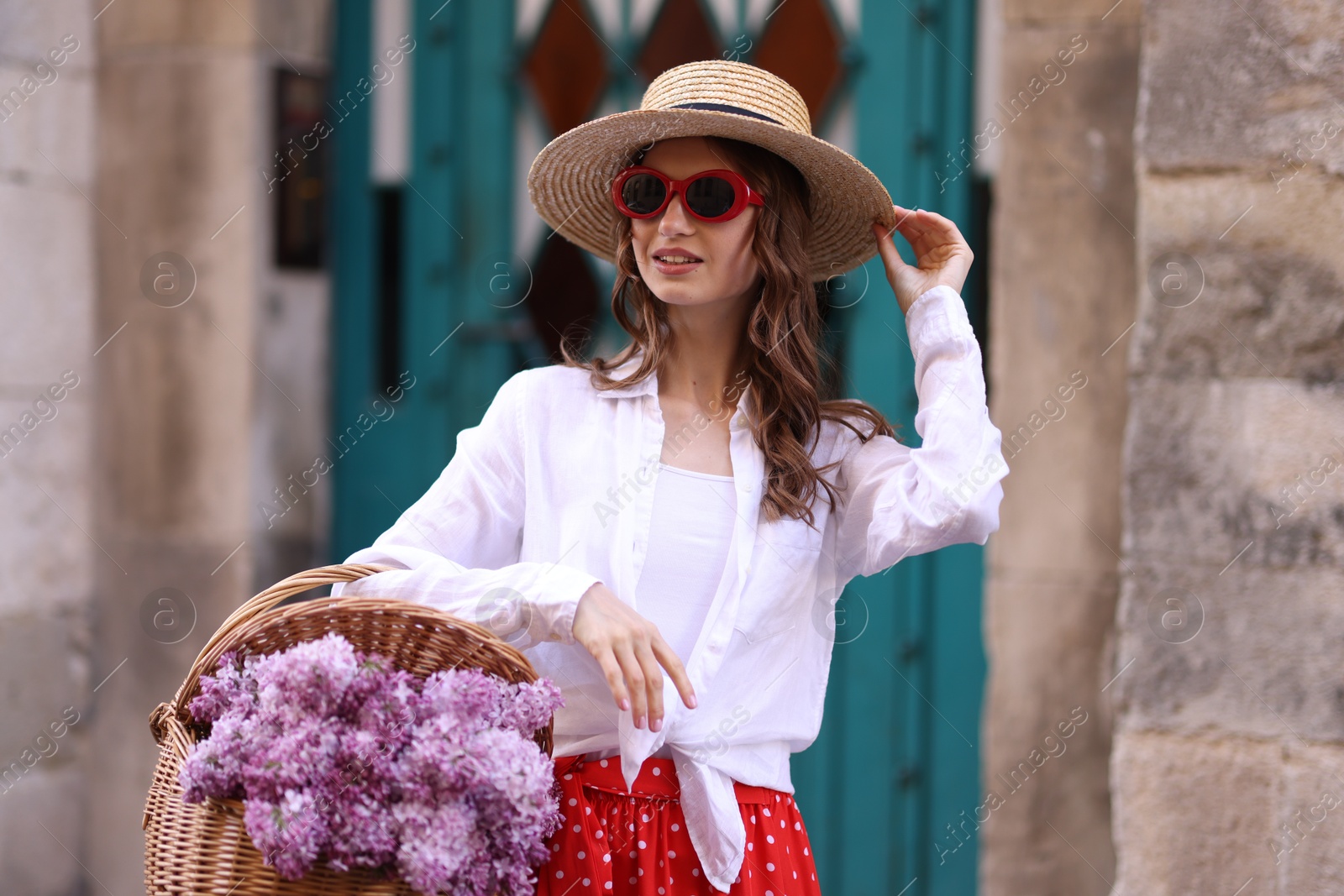 Smiling woman in hat and sunglasses with basket of lilac flowers near building outdoors Photo of Smiling woman in hat and sunglasses with basket of lilac flowers near building outdoors