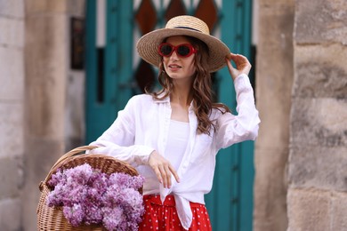 Photo of Smiling woman in hat and sunglasses with basket of lilac flowers near building outdoors