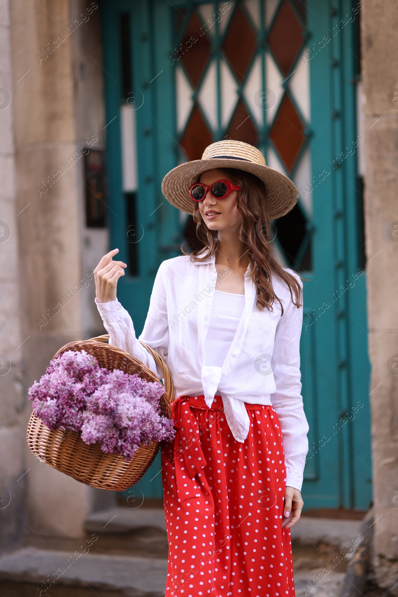 Smiling woman in hat and sunglasses with basket of lilac flowers near building outdoors Photo of Smiling woman in hat and sunglasses with basket of lilac flowers near building outdoors