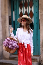 Photo of Smiling woman in hat and sunglasses with basket of lilac flowers near building outdoors
