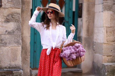 Photo of Beautiful woman in hat and sunglasses with basket of lilac flowers near building outdoors