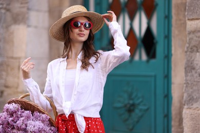 Photo of Beautiful woman in hat and sunglasses with basket of lilac flowers near building outdoors. Space for text
