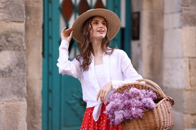 Photo of Smiling woman in hat with basket of lilac flowers near building outdoors