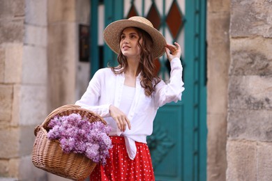 Photo of Smiling woman in hat with basket of lilac flowers near building outdoors
