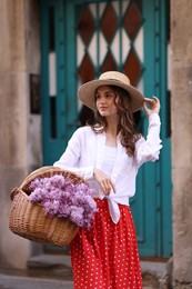 Photo of Beautiful woman in hat with basket of lilac flowers near building outdoors