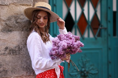 Photo of Beautiful woman in hat with bouquet of lilac flowers near building outdoors