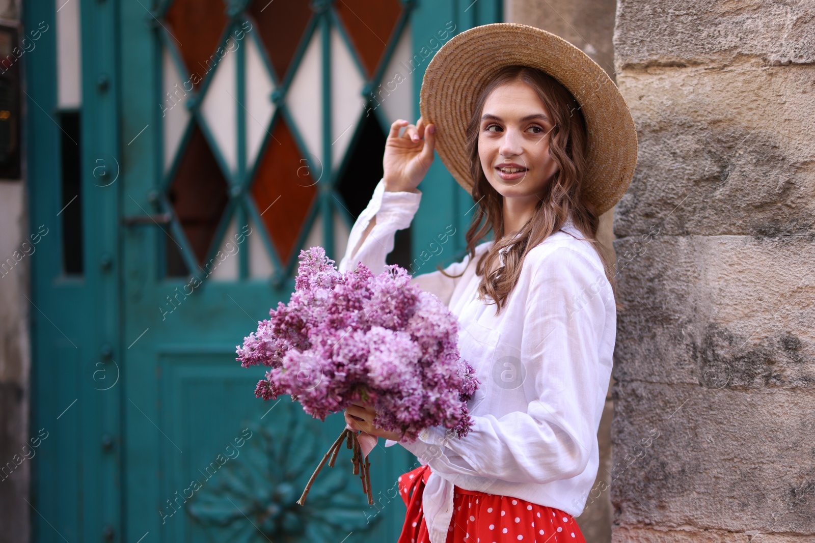Smiling woman in hat with bouquet of lilac flowers near building outdoors Photo of Smiling woman in hat with bouquet of lilac flowers near building outdoors