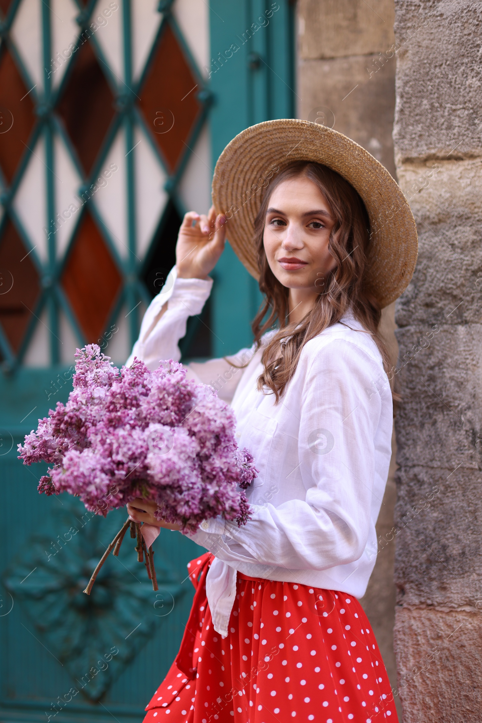 Beautiful woman in hat with bouquet of lilac flowers near building outdoors Photo of Beautiful woman in hat with bouquet of lilac flowers near building outdoors