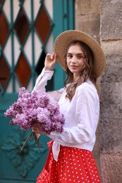 Photo of Beautiful woman in hat with bouquet of lilac flowers near building outdoors