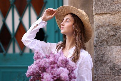 Photo of Beautiful woman in hat with bouquet of lilac flowers near building outdoors