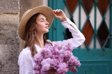 Photo of Beautiful woman in hat with bouquet of lilac flowers near building outdoors