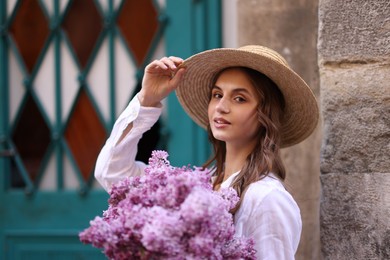 Photo of Beautiful woman in hat with bouquet of lilac flowers near building outdoors