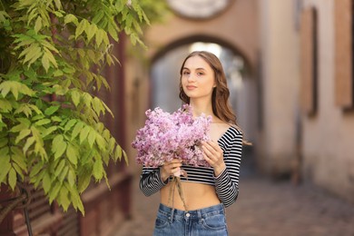 Photo of Beautiful woman with bouquet of lilac flowers near tree on city street